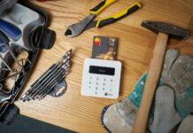 Construction payment - Overhead view of bright small plastic card in white contemporary wireless cash register surrounded by screwdriver set and hammer on big dirty gloves next to pliers